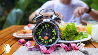 На изображении будильник на тарелке рядом с салатом и сантиметровой лентой — символика интервального голодания и контроля веса перед приемом пищи