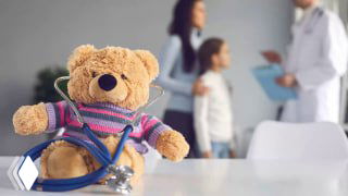Плюшевый медвежонок со стетоскопом на столе в кабинете; на фоне размыты врач и ребёнок — иллюстрация школьных жалоб.