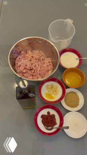 Крупный план ингредиентов для сирийской шаурмы: большая миска с мясом, рядом миски с йогуртом, лимоном, специями, маслом и солёными огурцами