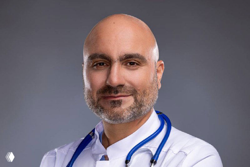 Крупный портрет врача-гастроэнтеролога в белом медицинском халате со стетоскопом на шее, спокойный деловой взгляд, нейтральный серый фон, студийная фотография.