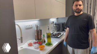 Мужчина стоит в кухне рядом со столом, на котором нарезанные овощи, специи и бутылка масла — сцена приготовления ингредиентов для баклажанной пасты