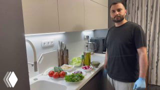 Человек на кухне у рабочей поверхности; на столе томаты, огурцы, пучки зелени, бутылки масла и подсушенный хлеб — подготовка к фаттушу.