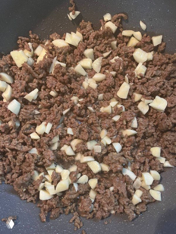 Обжаренный фарш с крупно нарезанным чесноком на сковороде — коричневая мясная масса и белые кусочки чеснока в процессе приготовления.