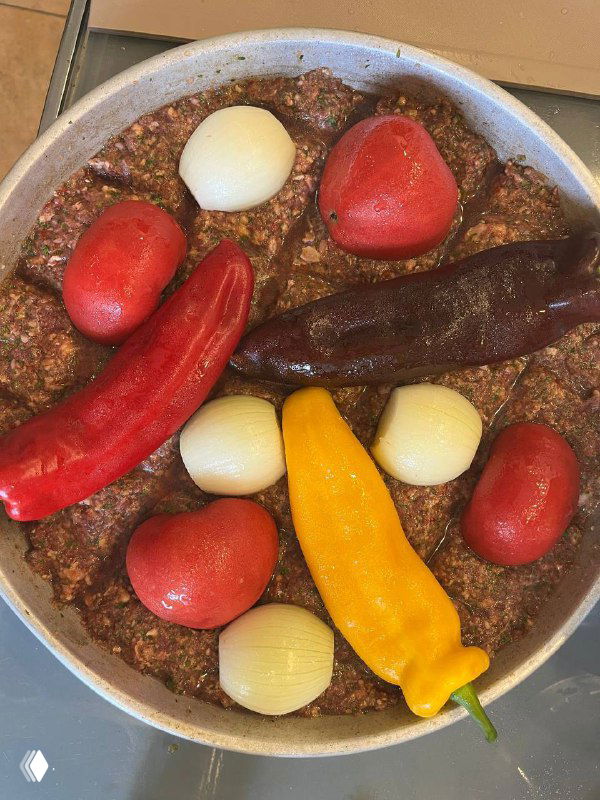 Собранный в противне фарш с поверхностью, на которой уложены целые помидоры, перцы рамиро и луковицы перед запеканием.