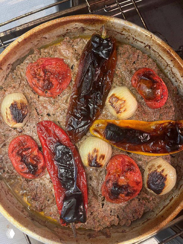 Противень после запекания: обугленные перцы, запечённые помидоры и лук на поверхности мясного пласта, видны карамелизация овощей.