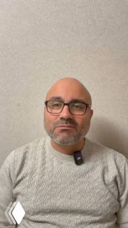 Портрет врача-гастроэнтеролога в очках и сером свитере на светлом нейтральном фоне; лицо смотрит в камеру, снимок статичный и информативный.