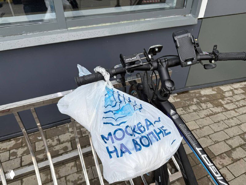 Полиэтиленовый пакет с логотипом «Москва на волне», подвешенный на руле велосипеда у фасада — брендовая упаковка доставки или самовывоза с морепродуктами