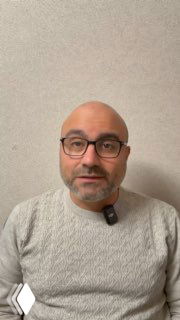 Портрет врача в крупном плане: Альмасри Али Мохамад в светлом свитере и очках, с петличным микрофоном на груди на нейтральном фоне, разговор в камеру.