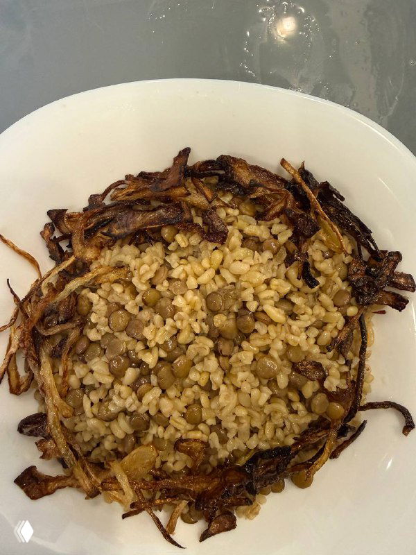 Тарелка мжадары — смесь булгура и чечевицы, сверху хрустящий карамелизированный лук; подана в белой миске, вид сверху.