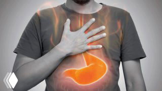 Мужчина держит руку на груди; графическое подсвечивание желудка и пищевода на фоне серого изображения, иллюстрация симптомов рефлюкса и изжоги.