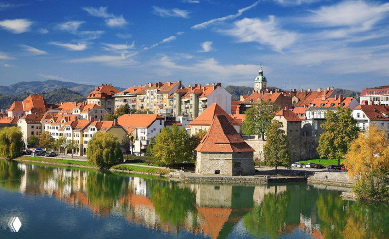 Панорамный вид словенского города у реки с красными крышами, зелёными холмами и ясным небом — иллюстрация жизни и пейзажа в Словении.