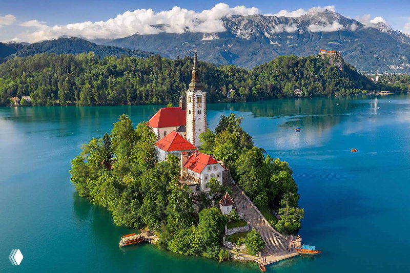 Панорамный вид на остров с церковью на озере Блед в Словении, окружённый зелёными деревьями, горами и спокойной водой.