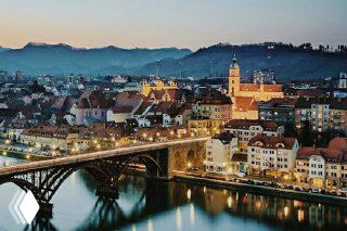 Вечерний городской пейзаж в Словении с освещённым мостом через реку и историческими домами на фоне холма — иллюстрация городского жилья и адресной регистрации.