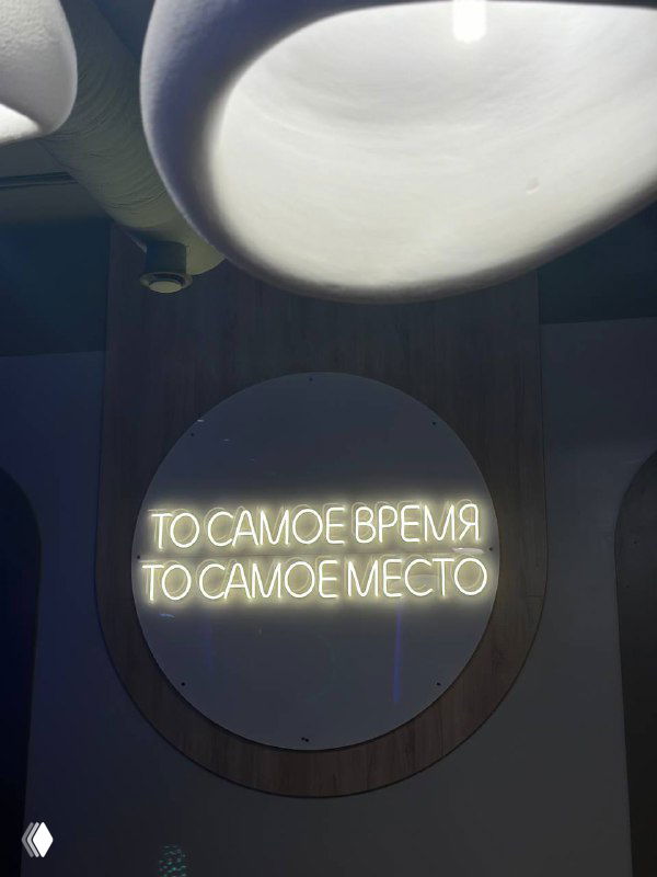 Неоновая вывеска в помещении мероприятия: круглая рама и надпись «ТО САМОЕ ВРЕМЯ ТО САМОЕ МЕСТО», интерьер площадки в тёмных тонах.