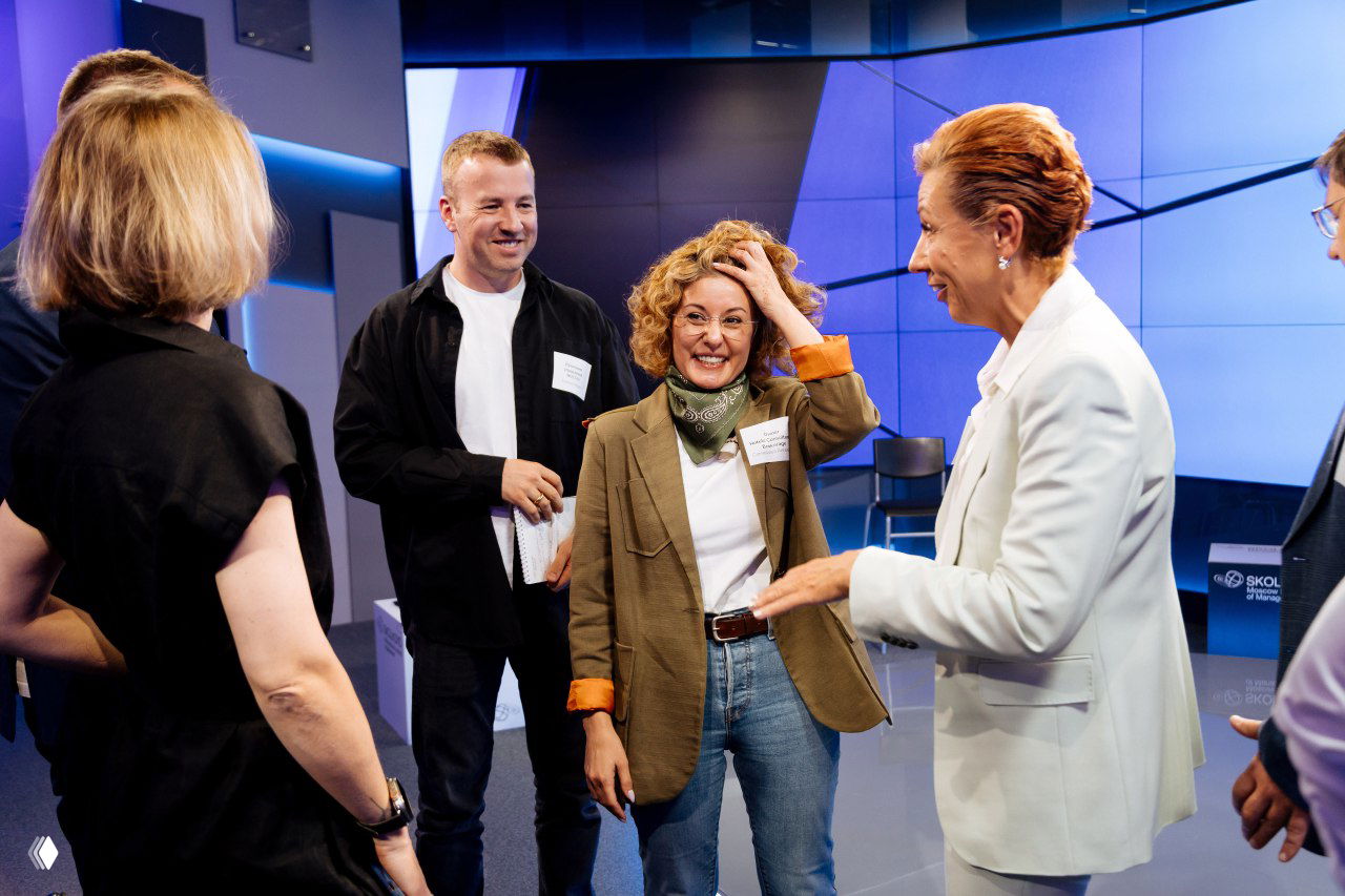 Ангелина Самадова общается с участниками в зале после сессии: несколько человек в деловой одежде, живая беседа и студийное освещение позади.