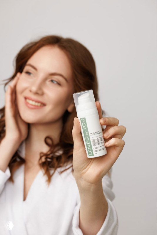 Молодая женщина держит флакон сыворотки Loved by You крупным планом, улыбается; видно лицо и упаковку продукта, акцент на уходе за кожей.