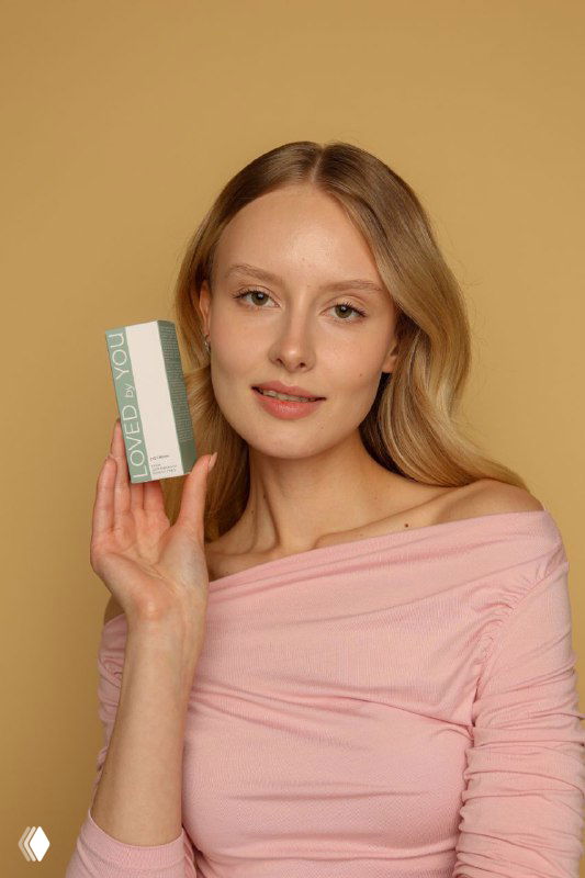 Девушка на светло-бежевом фоне держит коробочку косметики Loved by You; естественный макияж, акцент на упаковке и лице.