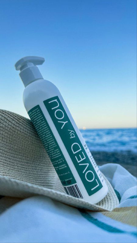 Флакон молочка Loved by You на пляжном фоне у моря: бутылка лежит на ткани у песка, четко видна этикетка бренда и летняя атмосфера ухода за кожей.