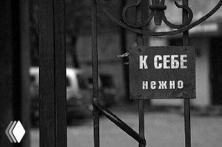 Чёрно-белая фотография металлической калитки с прикреплённой табличкой «К СЕБЕ нежно», размытый городской фон.