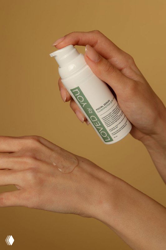 Крупный план рук наносит средство из флакона Loved by You: видно дозатор и текстуру средства на коже на бежевом фоне.