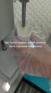 Процесс розлива косметического крема в баночки на производственной линии: металлическая насадка дозатора, рука в перчатке оператора и ряд заполненных ёмкостей