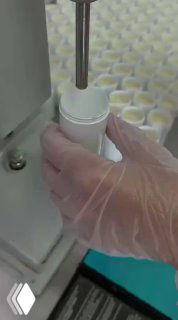 Крупный план производственной линии: в перчатках оператор держит флакон, дозатор наполняет крем — фасовка косметики и контроль качества на заводе.