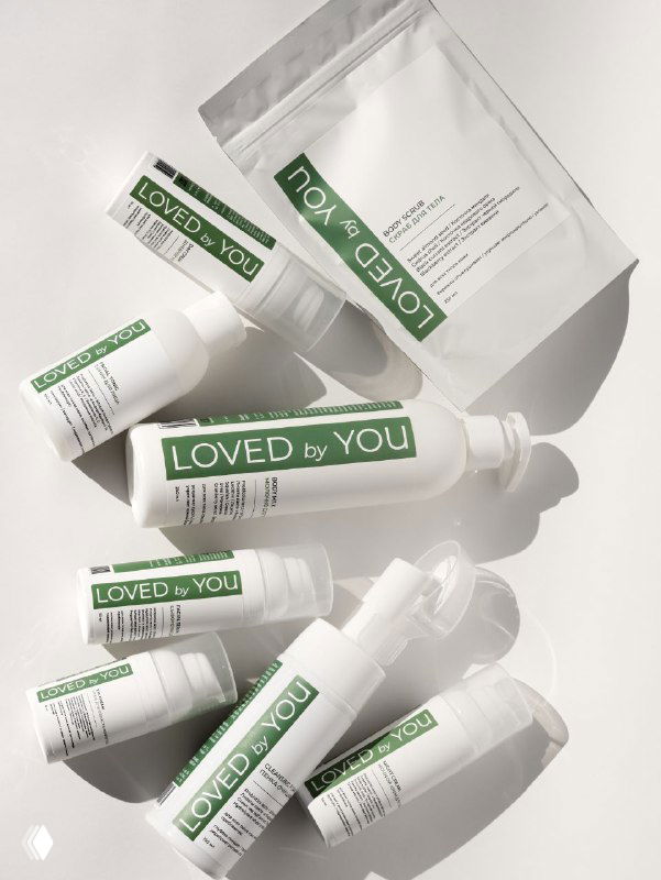 Плоская композиция белых баночек и тюбиков косметики Loved by You с зелёными этикетками на светлом фоне, видны тени.