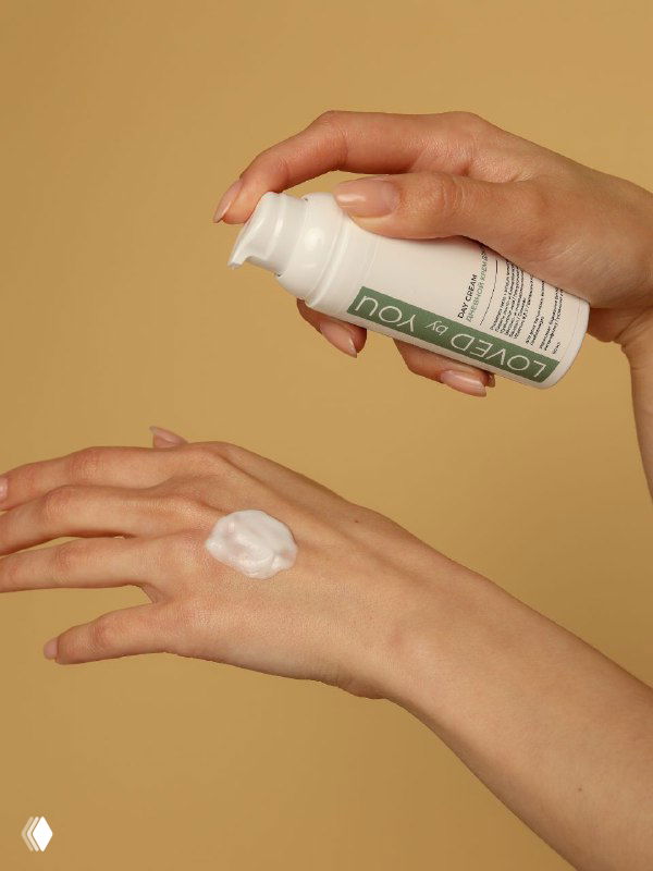 Руки наносят дневной крем Loved by You с дозатором на тыльную сторону кисти; фокус на текстуре крема и упаковке на бежевом фоне.