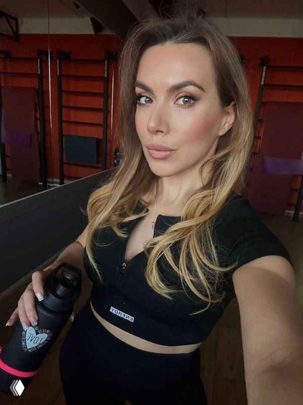 Портретная фотография женщины в спортивной одежде в тренажёрном зале, она держит бутылку для воды и улыбается в камеру; виден фон тренажёров.