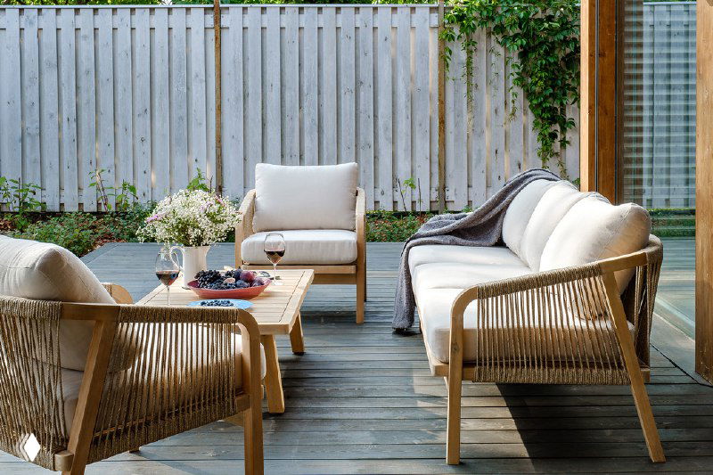 Вид под углом на комплект садовой мебели: диван и кресла с бежевыми подушками, деревянный стол и растительность вдоль забора.