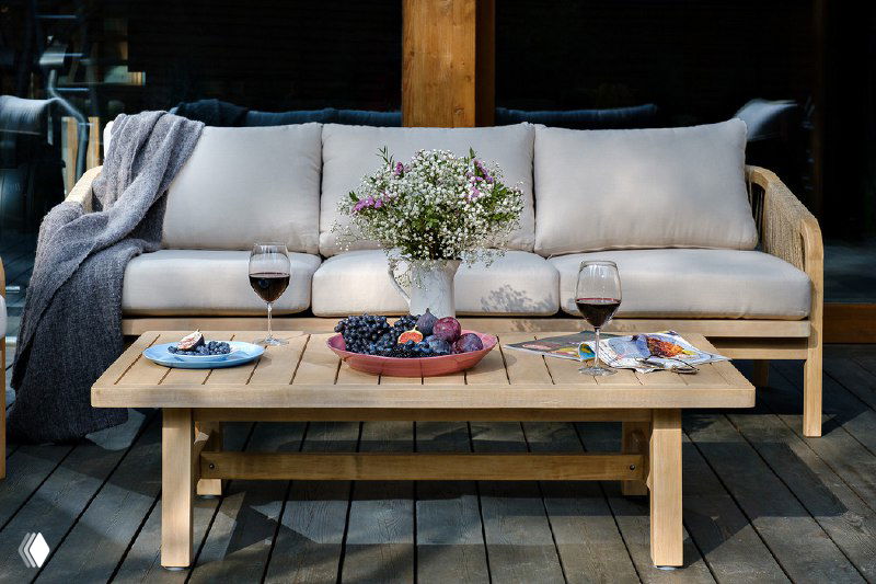 Светлый диван коллекции RIMINI с пледом и вазой с цветами на фоне кофейного столика и бокалов вина на террасе.