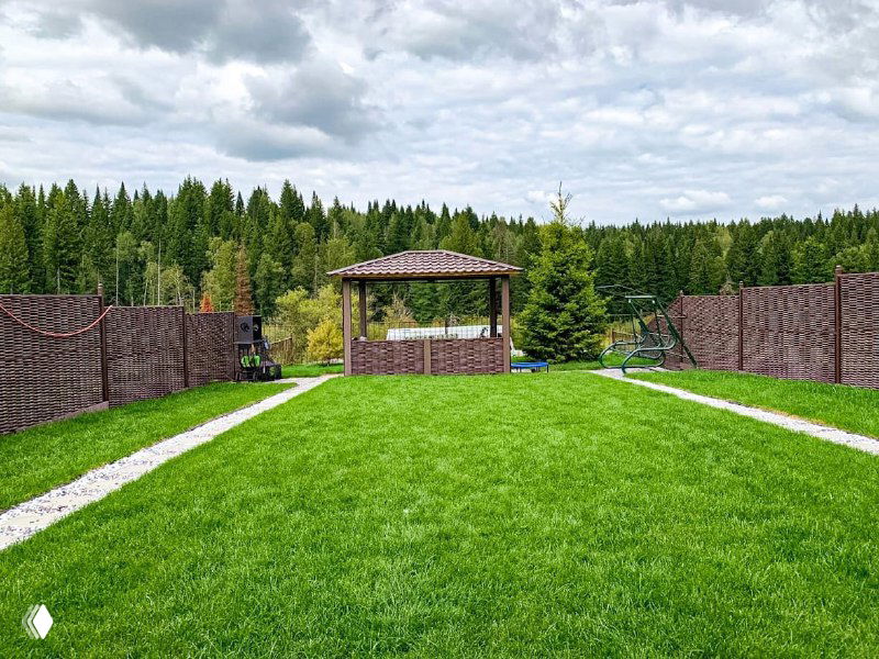 Прямой план широкого зелёного газона вокруг беседки; по бокам декоративная галька и забор, а на дальнем плане — густой хвойный лес.