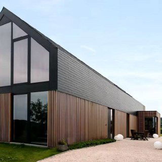 Современный фасад жилого дома, облицованный вертикальными брусковыми панелями из ДПК коричневого тона, показан с террасой и крупными окнами, подчёркивая внешний вид здания.