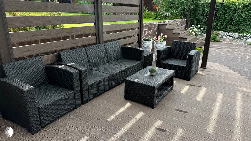 Уютная зона отдыха с комплектом пластиковой мебели из ротанга: диван, кресла и низкий столик на фоне декоративного ограждения участка.