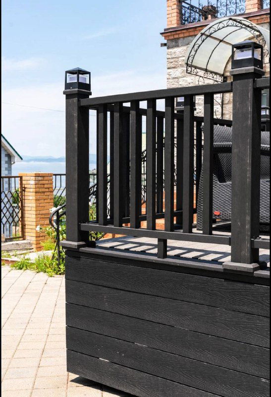 Угол ограждения и часть террасы днём: чёрная отделка, композитные панели в основании и декоративные перила, общий вид участка возле дома.