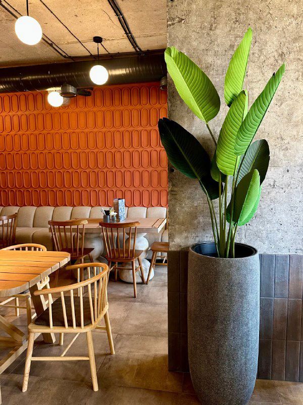 Высокий искусственный куст в большом сером вазоне на фоне оранжевой текстурной стены и столов ресторана, видна декорация зала.