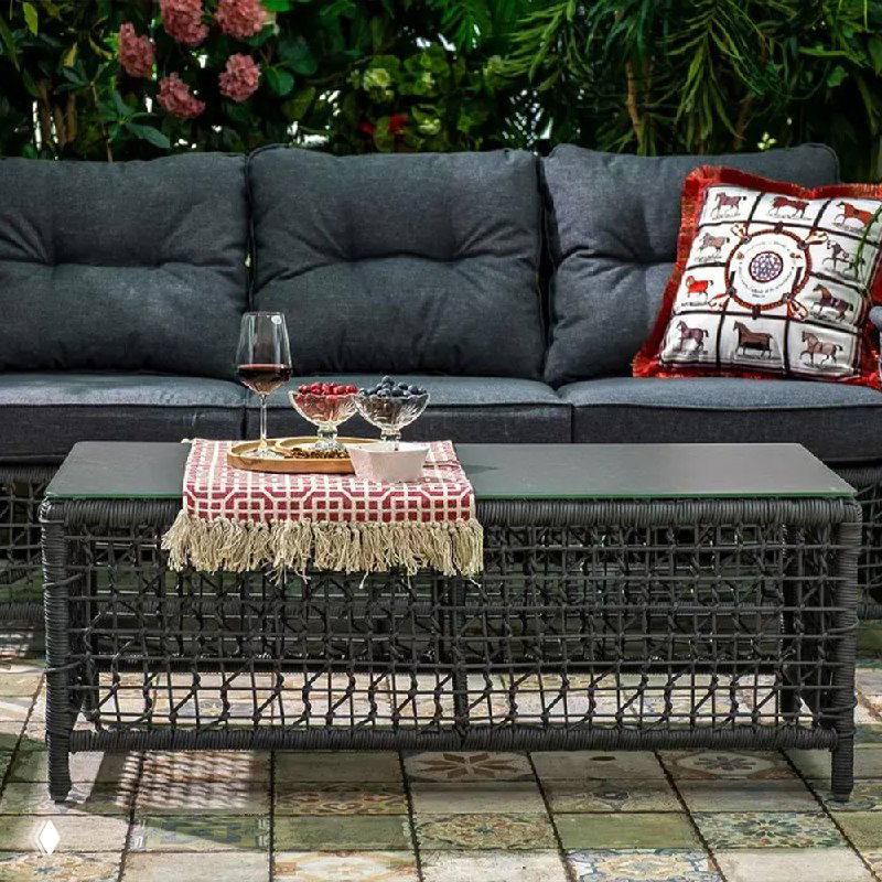 Угловой вид дивана и низкого стола LUCCA: тёмная обивка, керамическая столешница, боковая сервировка и зелёный растительный фон зимнего сада.