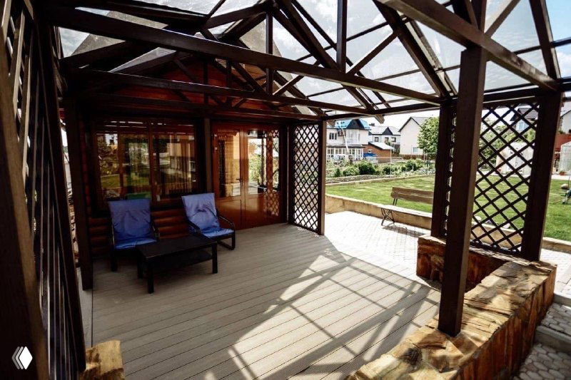 Вид внутри перголы из ДПК: настил, кресла и прозрачная крыша, солнечный день и ухоженный приусадебный участок; демонстрация эксплуатационных свойств материала.