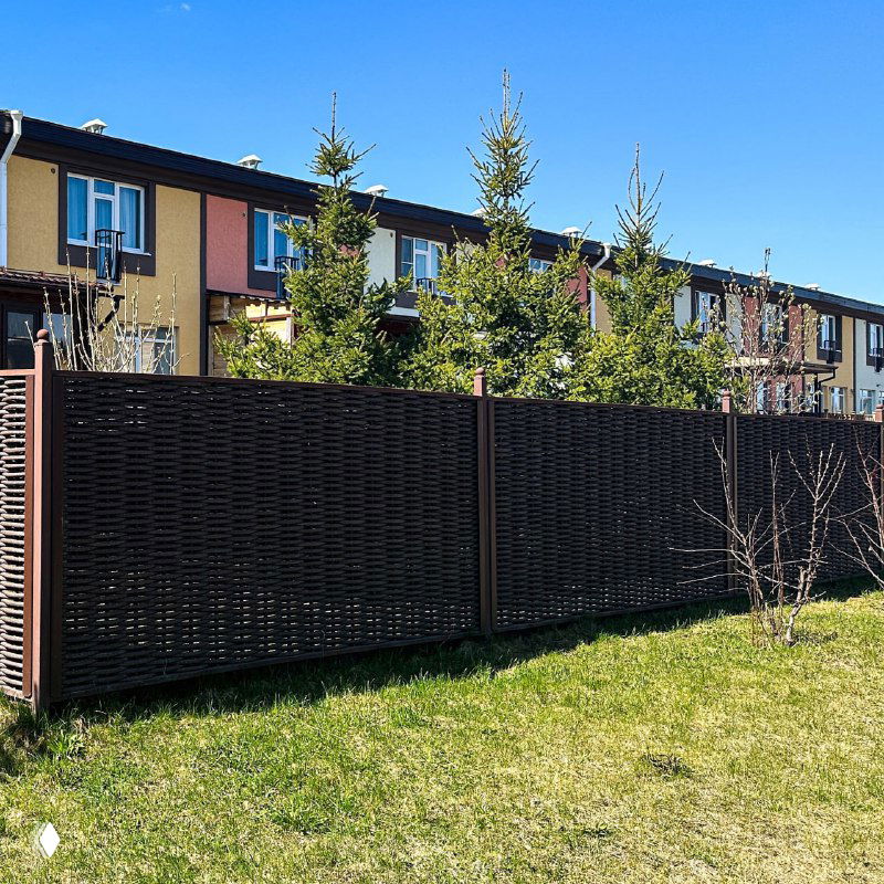 Длинная секция забора из полимерной лозы вдоль газона и ряд таунхаусов на заднем плане; наглядный пример зонирования участка и защиты от взглядов.