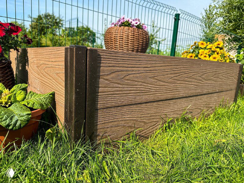 Крупный план уголка сборной грядки из ДПК: видна текстура панелей, стыки и трава с цветами вокруг.