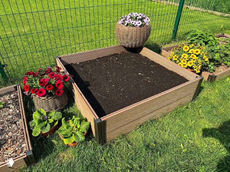 Сборная грядка из ДПК, заполненная тёмной плодородной почвой, окружена цветами и ухоженным газоном на участке.