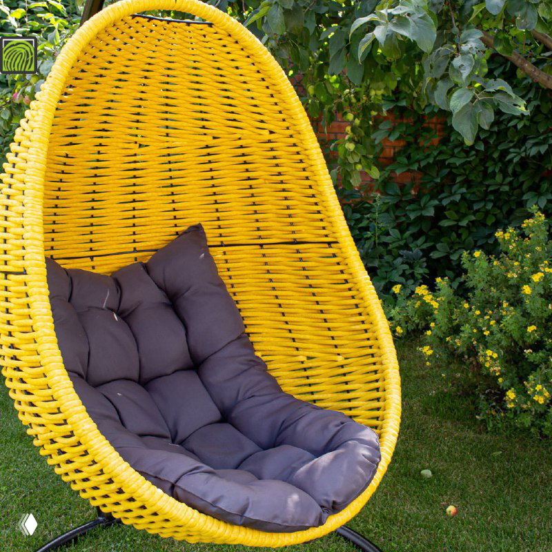 Жёлтое плетёное кресло‑кокон с тёмной мягкой подушкой, установленное в саду на фоне кустов и зелёной листвы, вид в полный рост.