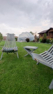 Два светло-серых складных дачных кресла и небольшой столик на зелёном газоне перед домом и теплицей под облачным небом, вид сбоку.