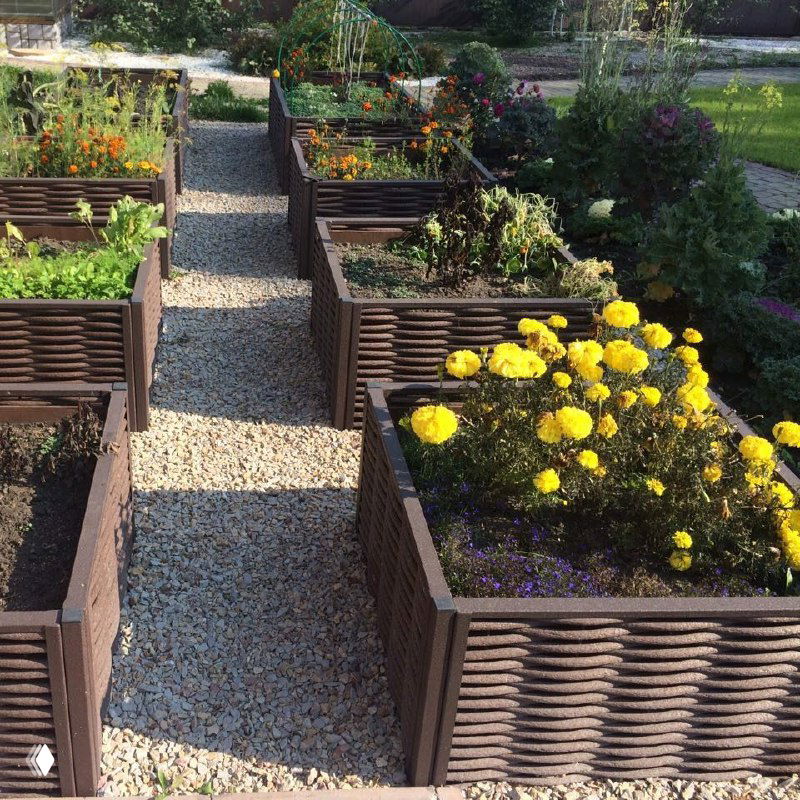 Несколько высоких грядок из плетёной полимерной лозы с цветами и овощами, аккуратные дорожки из гравия между грядками и зелёные клумбы.