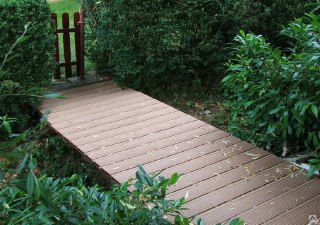 Дорожка из композитной террасной доски ДПК между кустами и зеленью — ровная нескользящая поверхность для прохода на участке.