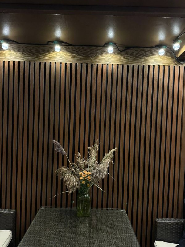 Тёмно-коричневые брусковые панели из ДПК с подсветкой сверху; внизу стеклянный стол и вазa с сухоцветами создают уютную сцену.