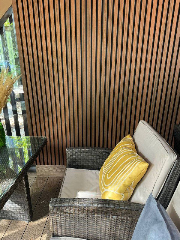 Крупный план стеновой панели из ДПК у зоны отдыха: ламели, текстура дерева и кресло с яркой декоративной подушкой.