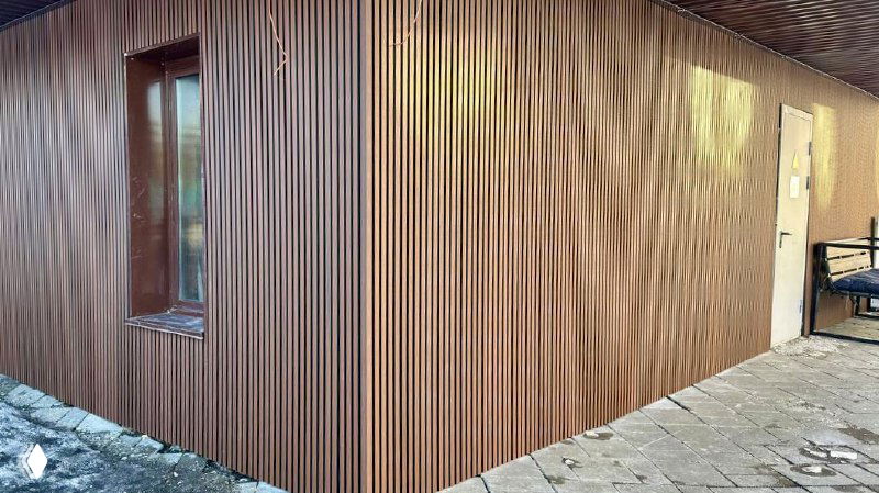 Крупный план угловой части фасада с вертикальными ламелями из ДПК; видно окно, фактуру панелей и часть лавочки на тротуаре в зимнем освещении.