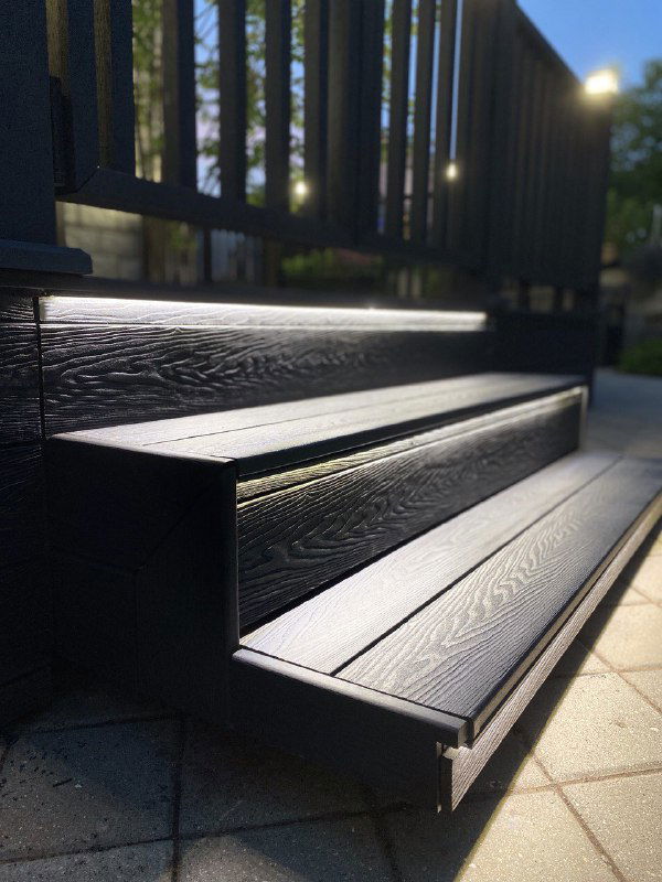 Снимок ступеней и настила террасы из тёмной террасной ДПК‑доски Polivan при вечерней подсветке, выделены линии ступеней и ворота.