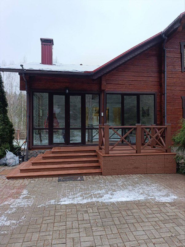 Общий вид красной террасы и входной зоны деревянного дома в зимне-весенний период; видна широкая лестница и настил из ДПК Polivan.